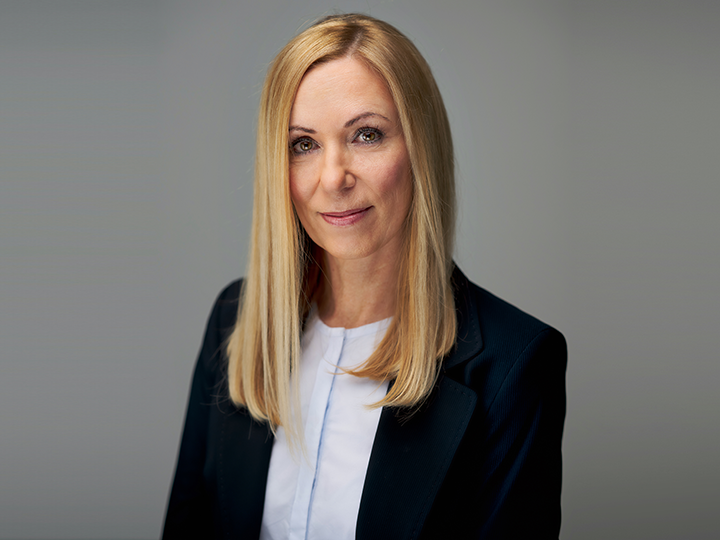 Una donna con lunghi capelli biondi indossa un blazer scuro sopra un top bianco. Sorride dolcemente e guarda la fotocamera su uno sfondo grigio semplice.