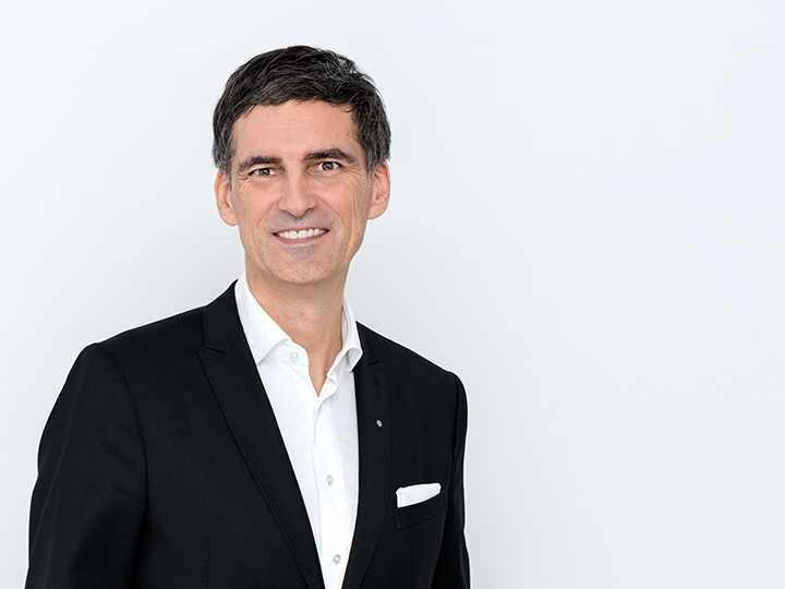 Un uomo in abito nero e camicia bianca è in piedi sorridendo di fronte a un semplice sfondo bianco. Ha i capelli corti e scuri e indossa un fazzoletto da taschino.