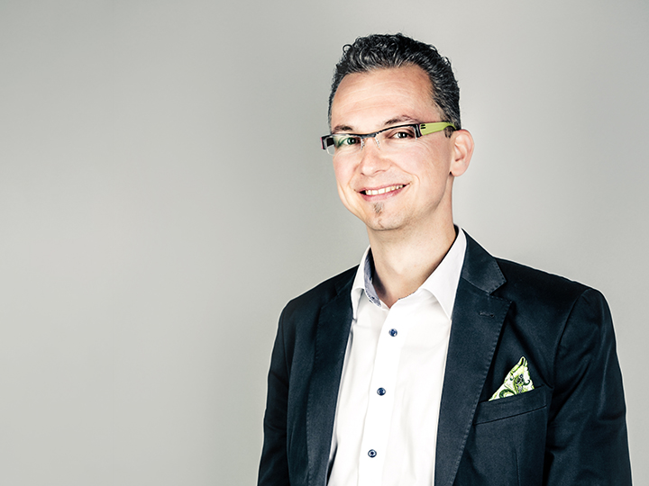 Un uomo con capelli corti scuri e occhiali sorride alla fotocamera. Indossa un blazer scuro sopra una camicia bianca con un fazzoletto da taschino fantasia. Lo sfondo è semplicemente grigio chiaro.