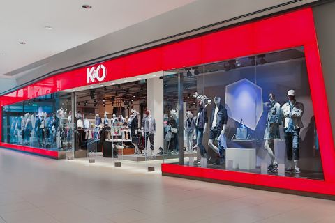 Vetrina con grande facciata in vetro rosso che espone manichini con abbigliamento casual. L'interno è ben visibile e mostra appendiabiti. Il logo del negozio mostra una "K&Ö" stilizzata.