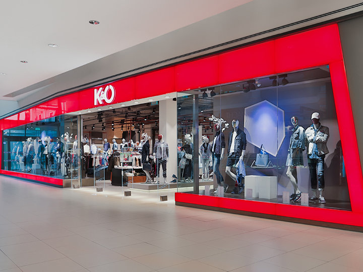 Vetrina con grande facciata in vetro rosso che espone manichini con abbigliamento casual. L'interno è ben visibile e mostra appendiabiti. Il logo del negozio mostra una "K&Ö" stilizzata.