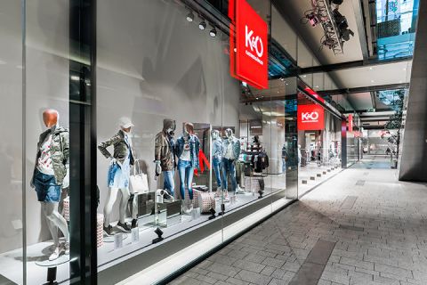 Una vetrina di un negozio di moda che espone manichini vestiti con abiti casual alla moda, tra cui giacche e pantaloncini di jeans. Il negozio ha grandi vetrate e si trova in un moderno centro commerciale con un'architettura elegante.