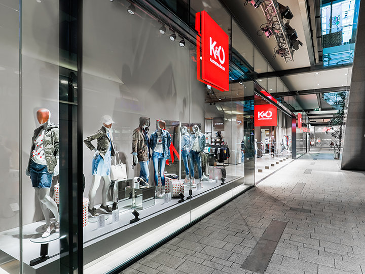 Una vetrina di un negozio di moda che espone manichini vestiti con abiti casual alla moda, tra cui giacche e pantaloncini di jeans. Il negozio ha grandi vetrate e si trova in un moderno centro commerciale con un'architettura elegante.