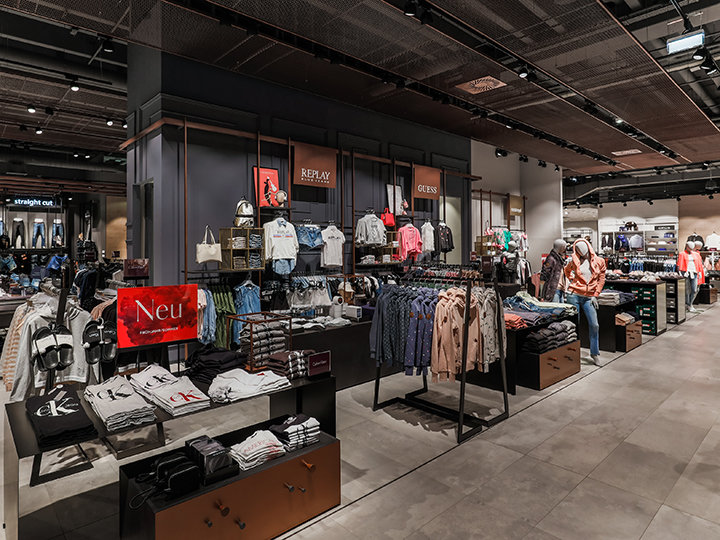 Un moderno negozio di abbigliamento con vari reparti che presentano ordinatamente camicie, giacche e accessori. Il negozio è caratterizzato da un'illuminazione brillante, esposizioni chiare e segnaletica del marchio, creando un ambiente di shopping elegante e invitante.