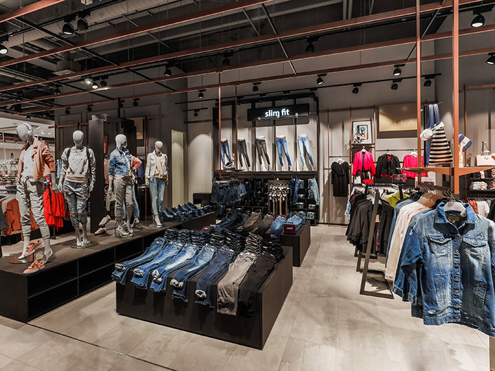 Interno di un negozio di abbigliamento con manichini che indossano completi di jeans. Tavoli espositivi presentano jeans piegati e scaffali espongono vari capi di abbigliamento. Il negozio ha un design moderno, con illuminazione industriale e un cartello con la scritta "Slim Fit".