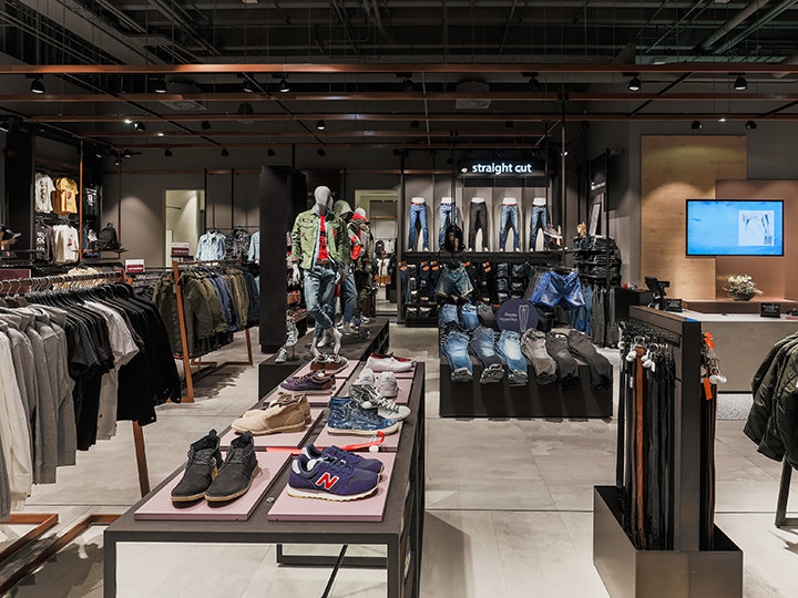 Un interno moderno di un negozio di abbigliamento con vari capi, tra cui camicie, giacche e jeans. Manichini espongono outfit, scarpe sono disposte sui tavoli e sullo sfondo si vede uno schermo digitale. L'insegna dice "taglio dritto".