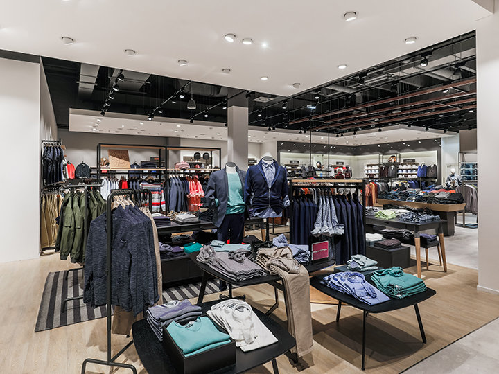 Un interno moderno di un negozio di abbigliamento con scaffali e tavoli ordinatamente disposti che espongono vari capi di vestiario come camicie, giacche e pantaloni. Lo spazio è ben illuminato da faretti e presenta un layout pulito e organizzato con tonalità neutre.