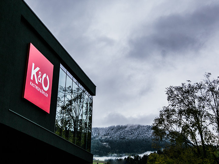 Un edificio moderno con un grande cartello rosso con la scritta "K&O Kastner & Öhler" è raffigurato di fronte a un cielo nuvoloso. Colline innevate e alberi frondosi circondano l'edificio, creando un contrasto tra elementi urbani e naturali.