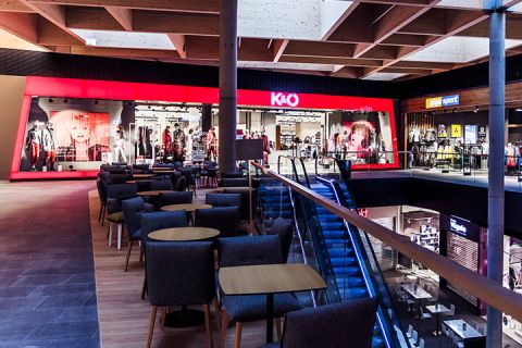 L'interno di un moderno centro commerciale con un negozio di abbigliamento illuminato di rosso chiamato K&O. Lo spazio comprende aree salotto con sedie e tavoli, una scala mobile e diverse altre vetrine sullo sfondo.