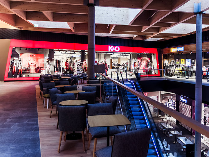 L'interno di un moderno centro commerciale con un negozio di abbigliamento illuminato di rosso chiamato K&O. Lo spazio comprende aree salotto con sedie e tavoli, una scala mobile e diverse altre vetrine sullo sfondo.