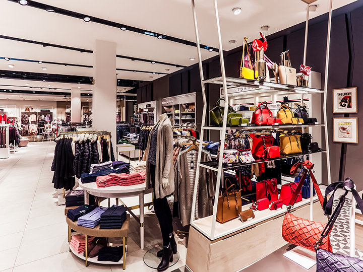 Un negozio di abbigliamento e accessori con scaffali pieni di borse colorate, sciarpe e vestiti piegati ordinatamente. Manichini indossano abiti alla moda. Il negozio è ben illuminato e arredato in stile moderno.