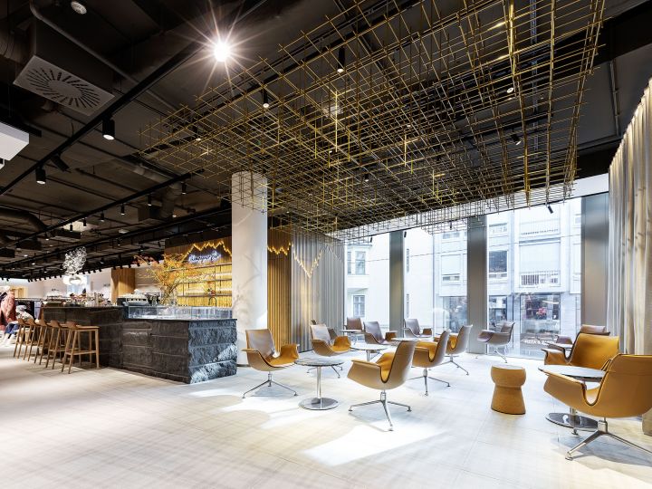 Moderna area lounge con eleganti sedie marroni e tavolini su un pavimento chiaro. Il soffitto presenta una struttura geometrica dorata. Grandi finestre lasciano entrare molta luce naturale e sullo sfondo si intravede un bar con sgabelli.