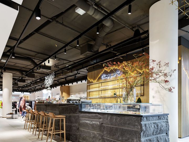 Interni moderni di un caffè con bancone in pietra, sgabelli da bar in legno e piante ornamentali. Il soffitto è scuro e dotato di illuminazione su binario, sullo sfondo si intravede un'area salotto con persone. Un'installazione geometrica a soffitto in oro aggiunge un tocco di stile all'ambiente.