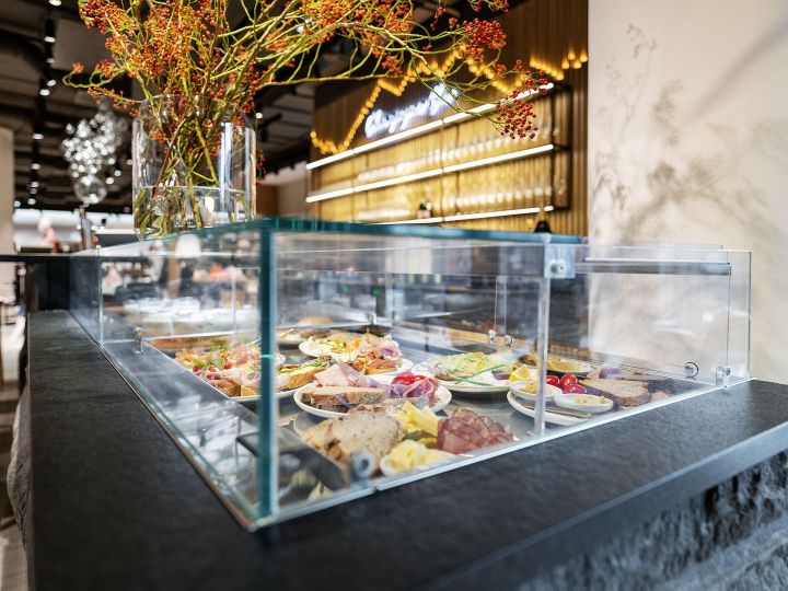 Vetrina in vetro in un ristorante moderno che espone vari piatti su piatti, tra cui carne, formaggio e verdure. Sopra sono disposti rami decorativi con bacche rosse. Parete in legno con segnaletica illuminata sullo sfondo.