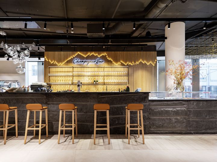 Un moderno champagne bar con un elegante bancone in pietra e cinque sgabelli marroni. Dietro il bancone, bicchieri e bottiglie sono esposti su scaffali. Un'insegna al neon recita "Champagne Bar". Una lampada decorativa e una composizione floreale aggiungono eleganza all'ambiente.