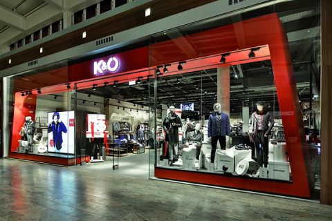 Vetrine di K&Ö in un centro commerciale con manichini che indossano vari stili di abbigliamento. L'esposizione comprende giacche, camicie e accessori sotto una luce intensa, all'interno si possono vedere altri capi di abbigliamento.