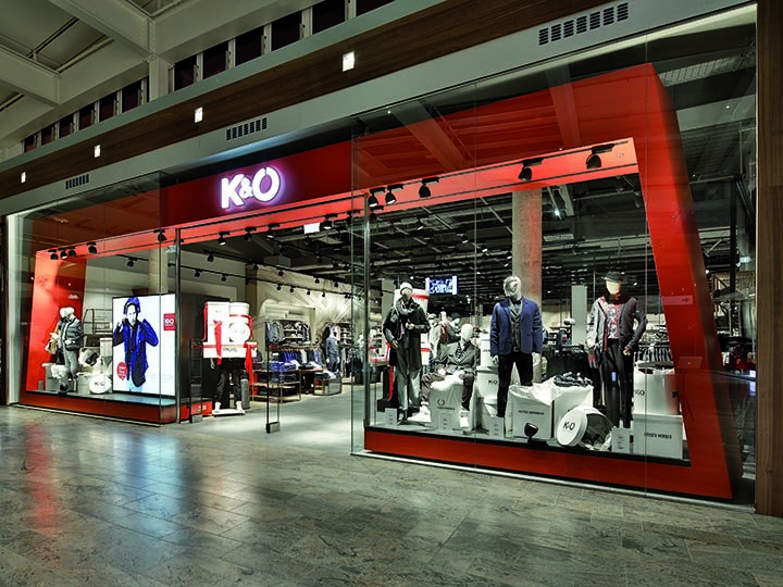 Vetrine di K&Ö in un centro commerciale con manichini che indossano vari stili di abbigliamento. L'esposizione comprende giacche, camicie e accessori sotto una luce intensa, all'interno si possono vedere altri capi di abbigliamento.