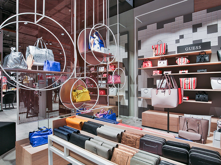 Un'elegante esposizione di borse in vari colori e stili presenta scaffali e appendini circolari. L'interno luminoso e moderno del negozio è caratterizzato da un'insegna Guess ben visibile tra le borse disposte ordinatamente.