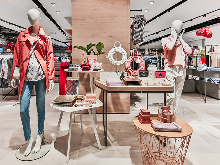 Un'elegante vetrina per un negozio di abbigliamento con manichini che indossano abiti primaverili casual. Accessori come borse e gioielli sono esposti sui tavoli. Il negozio è ben illuminato e ben organizzato, creando un ambiente di shopping moderno e invitante.