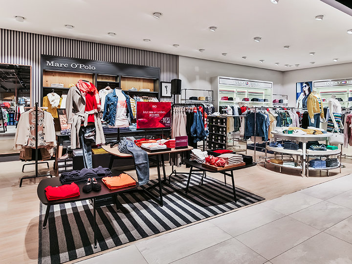 Un moderno negozio di abbigliamento con scaffali e espositori ordinatamente disposti con camicie, giacche e pantaloni colorati. Il negozio è ben illuminato e sul pavimento c'è un tappeto a righe. Sullo sfondo si vedono marchi come "Mare O'Polo".