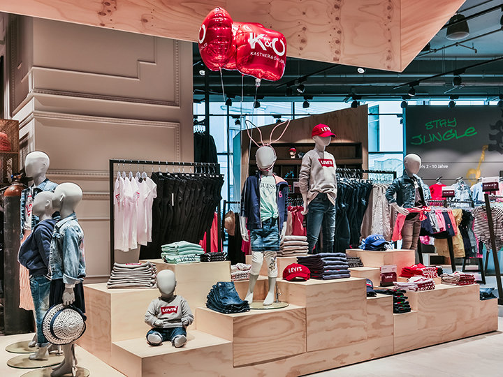 Vetrina di un negozio di abbigliamento per bambini con manichini vestiti in modo casual, tra cui jeans e camicie rosse. Gli scaffali sono pieni di vestiti piegati e accessori. Palloncini rossi pendono sopra e sullo sfondo un'insegna al neon recita "STAY JUNGLE".