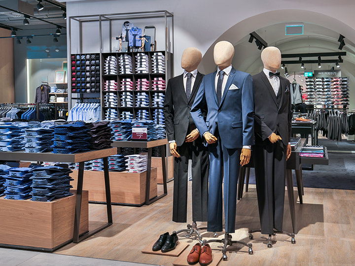 Una vetrina di un negozio di abbigliamento con tre manichini in abiti formali. Nell'ambiente circostante si possono notare camicie e accessori ben impilati. Il negozio ha un interno moderno con illuminazione soffusa e merce ben ordinata.