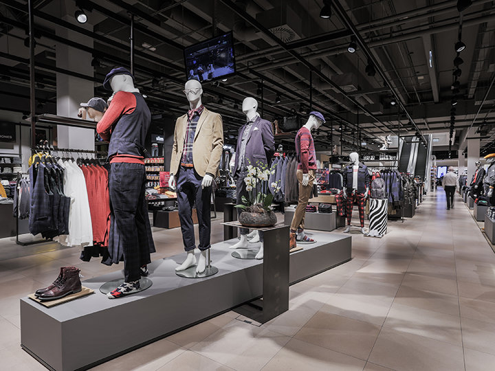 Una vetrina di un negozio di abbigliamento con manichini che indossano vari abiti casual e da sera. Il negozio è spazioso, sullo sfondo si vedono appendiabiti con vestiti e accessori sotto una luce intensa. Una persona sta camminando nel corridoio.