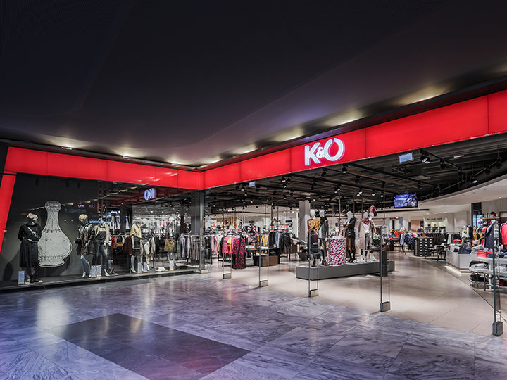 Vetrine di un negozio di moda K&O con manichini che espongono vari outfit dietro ampie vetrate. Il negozio presenta un design elegante e moderno e un'insegna rosso brillante con il logo K&O ben visibile sopra l'ingresso.