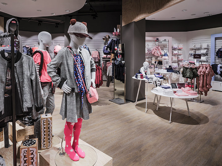 Un negozio di abbigliamento per bambini con manichini che indossano abiti colorati, tra cui un cappello e leggings rosa. Vari capi di abbigliamento e accessori sono esposti su scaffali e stand. Il pavimento è in legno e il negozio ha un arredamento interno moderno.