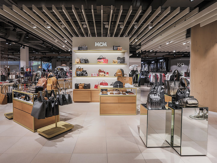 Un moderno interno di negozio che espone una varietà di borse di lusso su espositori in legno e vetro. Altre borse sono visibili sugli scaffali a parete. Il soffitto presenta un design geometrico e l'illuminazione è brillante, mettendo in risalto la merce.