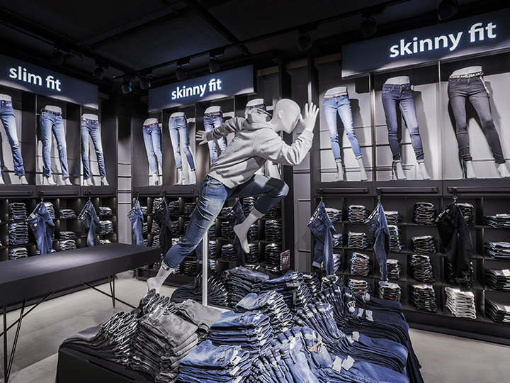 In una vetrina sono esposti diversi stili di jeans, tra cui slim fit e skinny fit. Sopra un tavolo c'è un manichino in posa dinamica, che indossa jeans e una felpa con cappuccio. Sullo sfondo, gli scaffali sono pieni di jeans piegati.
