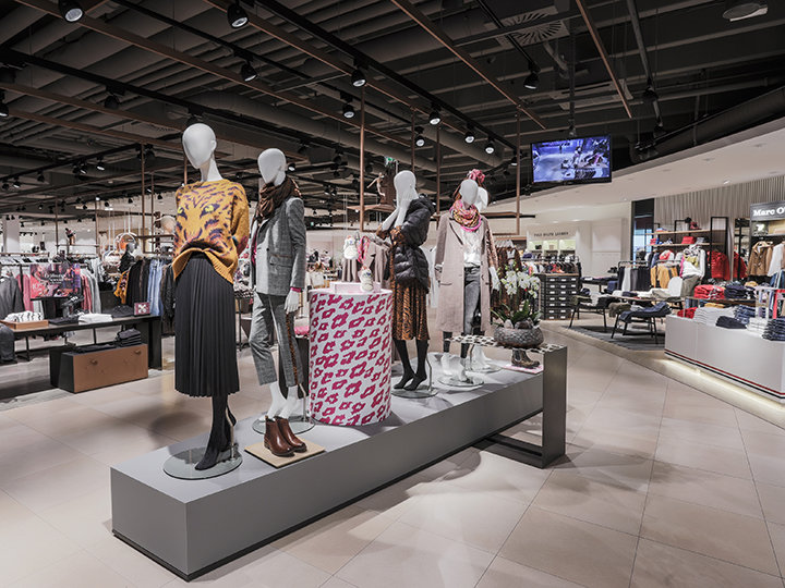 Un elegante interno di un negozio di abbigliamento con manichini che indossano abiti alla moda. Il negozio ha un arredamento moderno, appendiabiti e una grande scatola a motivi. L'illuminazione del soffitto mette in risalto diverse esposizioni e uno schermo è visibile sullo sfondo.