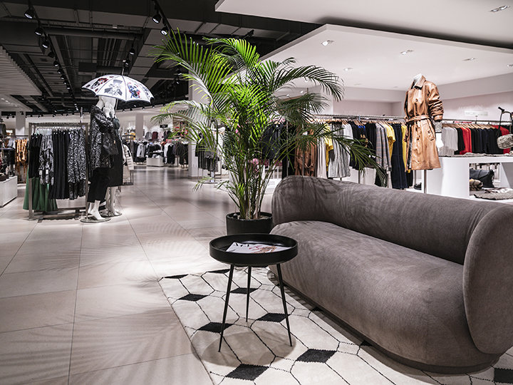 Un interno moderno di un negozio di abbigliamento con appendiabiti, tra cui cappotti e giacche. In primo piano ci sono un divano grigio e un tavolo rotondo nero accanto a una pianta in vaso. Sullo sfondo, un manichino tiene un ombrello.