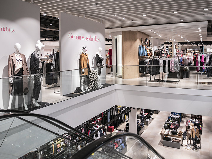 Un interno moderno di un negozio di abbigliamento con manichini in abiti casual esposti su una piattaforma. Appendiabiti e scaffali sono ordinatamente disposti su due piani, visibili dalla scala mobile in primo piano.