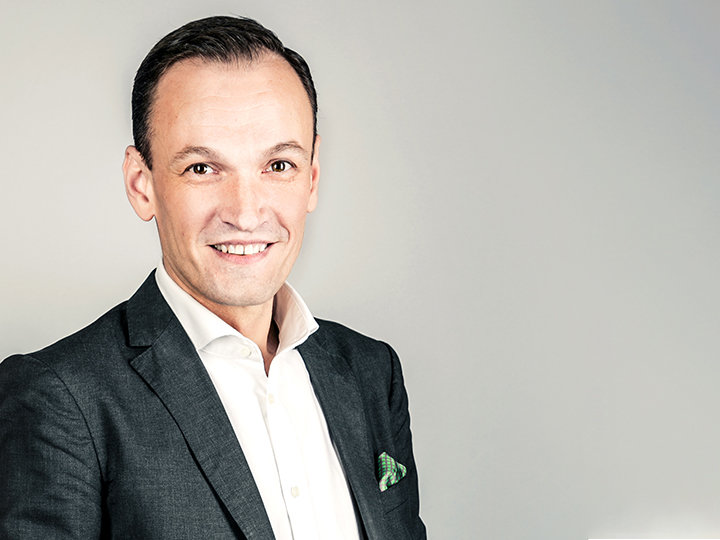 Un uomo in abito scuro e camicia bianca sorride davanti a uno sfondo grigio chiaro. Ha i capelli corti e un fazzoletto da taschino nella giacca.