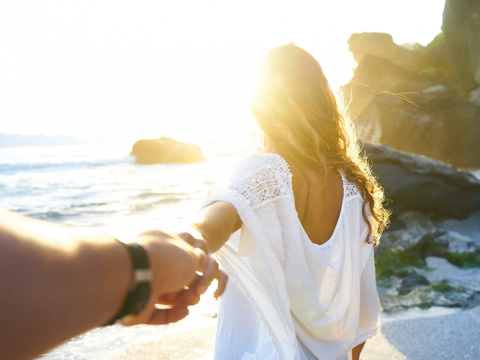 Una persona in abito bianco cammina lungo una spiaggia soleggiata tenendo la mano di un'altra persona. Sullo sfondo si vedono il mare e grandi rocce, e la luce del sole crea un'atmosfera calda e luminosa.
