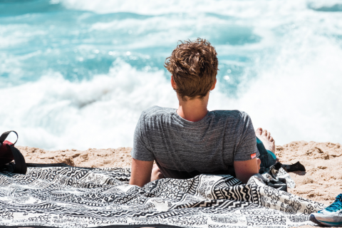 Una persona con i capelli corti e una maglietta grigia è sdraiata su un asciugamano a motivi sulla spiaggia. Guarda il mare, sullo sfondo si infrangono le onde.