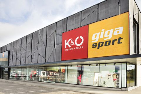 Vista esterna di un edificio moderno con grandi insegne per "Kastner & Öhler" in rosso e "Giga Sport" in giallo. Al piano terra si vedono ampie vetrine con articoli sportivi e di moda. Il cielo è coperto.