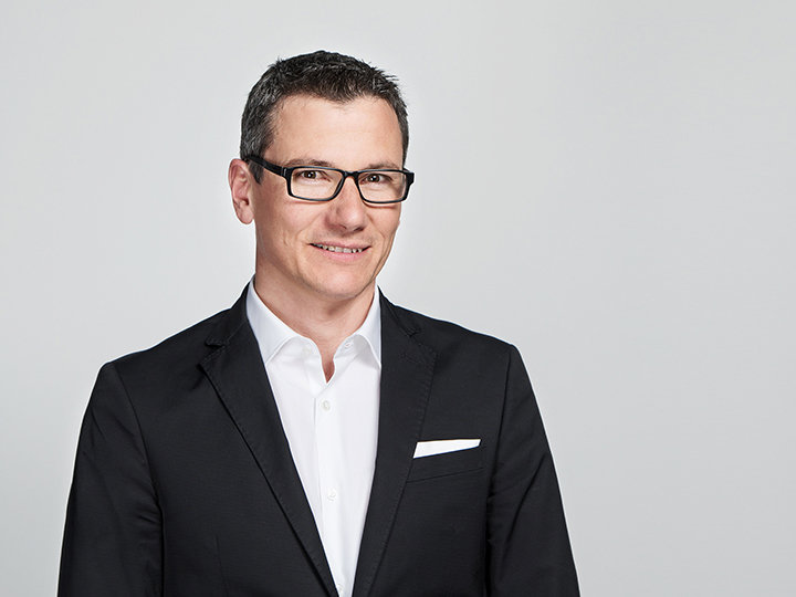 Una persona con occhiali, vestito nero e camicia bianca sorride alla fotocamera. Lo sfondo è semplicemente grigio chiaro.