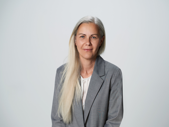 Una persona con lunghi capelli biondi, un blazer grigio chiaro e una camicetta bianca è in piedi davanti a uno sfondo semplice grigio chiaro. Ha un'espressione neutra e guarda la telecamera.