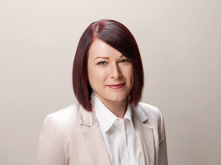 Una persona con capelli rosso scuro lunghi fino alle spalle, un blazer chiaro e una camicia bianca sorride alla telecamera su uno sfondo neutro.