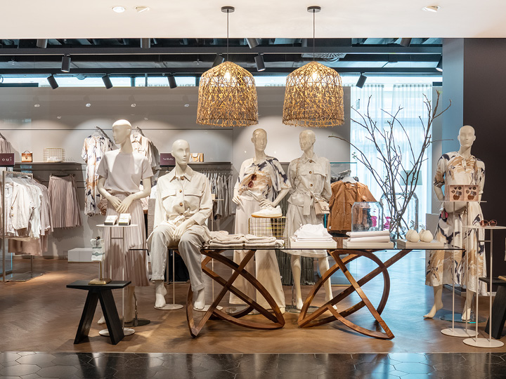 In un negozio di abbigliamento, diversi manichini sono esposti con abiti neutri e alla moda. Sono circondati da un'illuminazione soffusa, un arredamento moderno e capi di abbigliamento ordinatamente disposti su stand e tavoli. Sopra di loro pendono lampade a sospensione intrecciate.