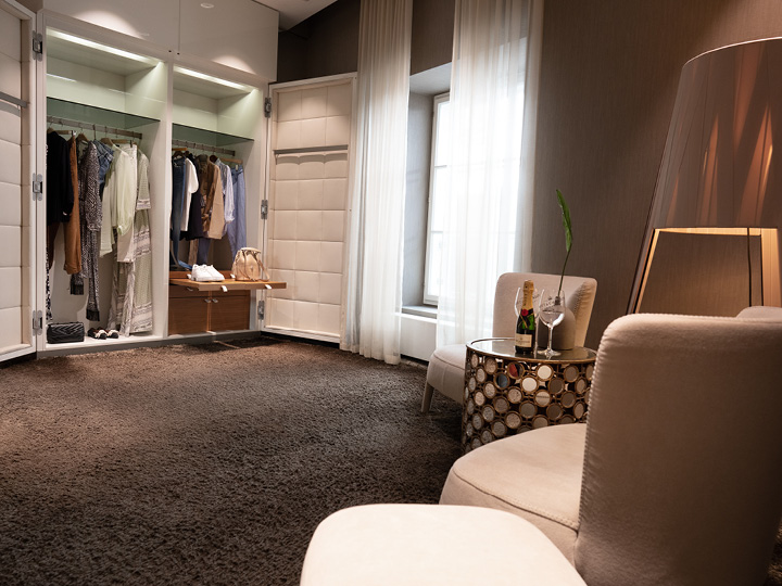 Una stanza moderna ed elegante con una cabina armadio a vista che espone abiti e una borsa. Due comode sedie sono posizionate di fronte a un tavolo rotondo con una lampada decorativa. Una bottiglia di champagne e bicchieri sono sul tavolo e la luce filtra attraverso le tende trasparenti.