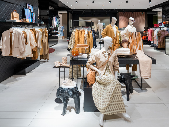 Interni di un negozio di abbigliamento con vestiti in tonalità neutre su appendiabiti e manichini. In esposizione camicie, giacche, vestiti e accessori come borse e cappelli. Illuminazione chiara, con un layout moderno e pulito.