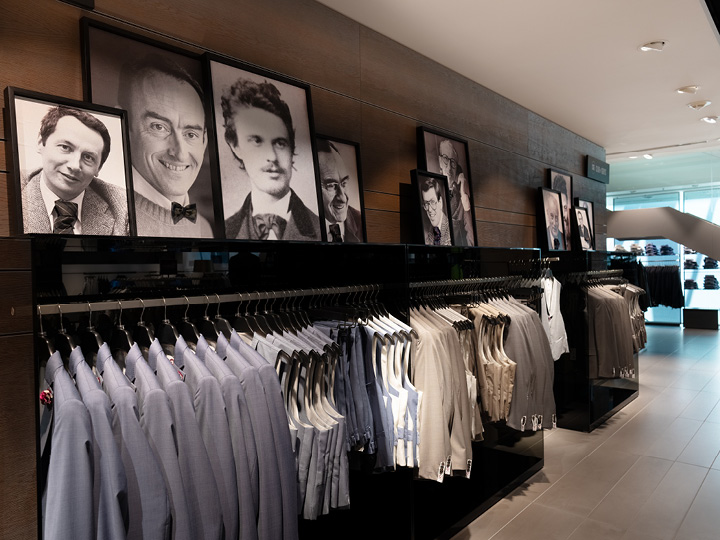In un negozio di abbigliamento, abiti in varie tonalità di blu e beige sono appesi ordinatamente. Sopra gli abiti, ritratti in bianco e nero di diversi uomini sono appesi a una parete di legno.