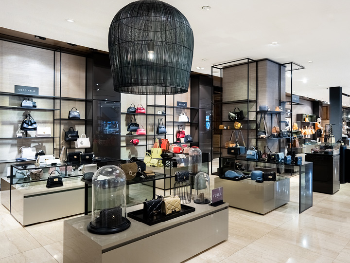 Un negozio di lusso espone borse firmate su eleganti scaffali e banconi. Lo spazio è dotato di illuminazione moderna, toni neutri e un grande lampadario nero che crea un'elegante atmosfera per lo shopping.