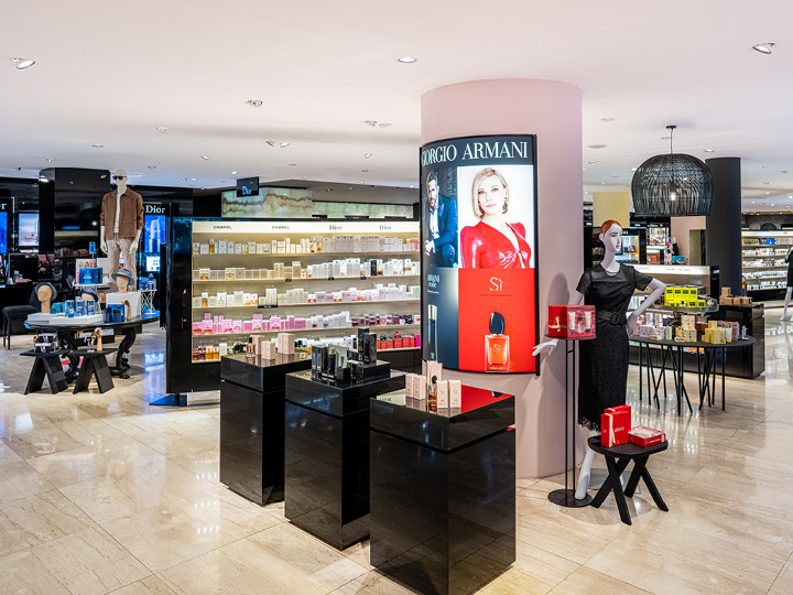 Un grande magazzino di lusso espone vari marchi di profumi, tra cui Giorgio Armani. Luci brillanti illuminano moderni espositori neri pieni di prodotti. Sullo sfondo, scaffali pieni di altri articoli di bellezza.