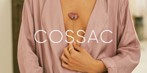Una persona in una veste di seta rosa chiaro tiene tra le dita uno stelo di fiori rosa vicino al petto. La parola "COSSAC" è sovrapposta all'immagine in caratteri bianchi.