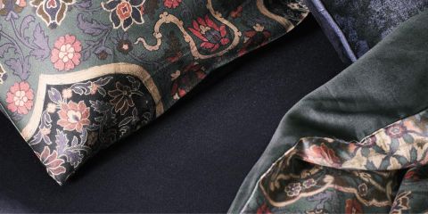 Primo piano di un letto con cuscini scuri a motivi floreali e una coperta abbinata. Il tessuto presenta intricati disegni nei toni del rosso, rosa e verde, che risaltano su uno sfondo scuro. Il tessuto appare soffice e morbido.