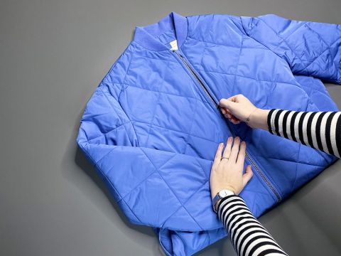 Una persona con una maglia a maniche lunghe a righe chiude la zip di una giacca imbottita blu appoggiata su una superficie grigia.